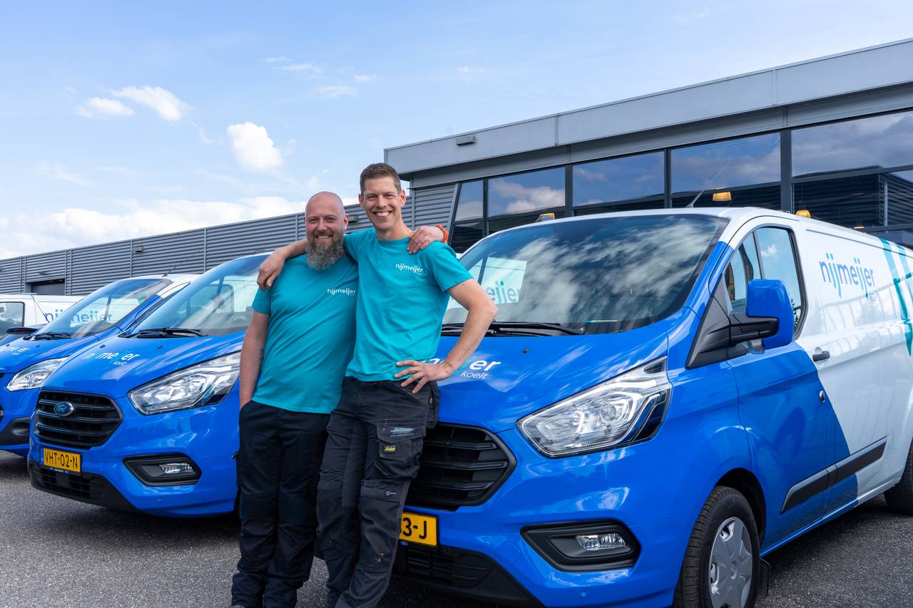 Twee medewerkers die een arm om elkaar heen geslagen hebben, leunen tegen de bedrijfsbus.